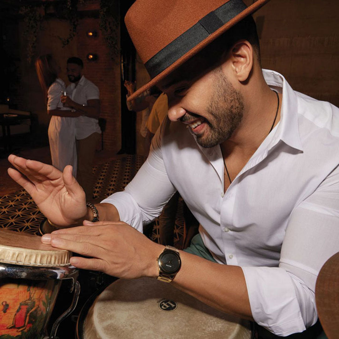 A smiling man in a brown hat and white shirt plays a conga drum, surrounded by modern design elements and patterned flooring. In the background, people holding drinks admire his rhythm, while he discreetly displays a Bulova Millennia Quartz 41mm Watch on his wrist.