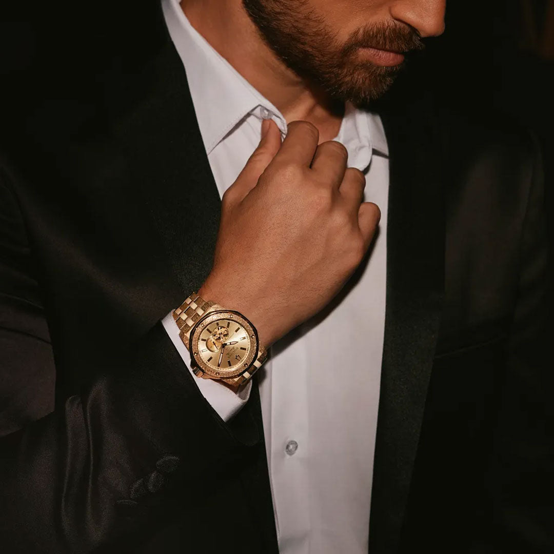 A man in a black suit and white shirt adjusts his collar, highlighting the Bulova Marine Star Marc Anthony Automatic 44mm Watch on his wrist. The dark background accentuates both the elegant Bulova timepiece and his sharp attire.