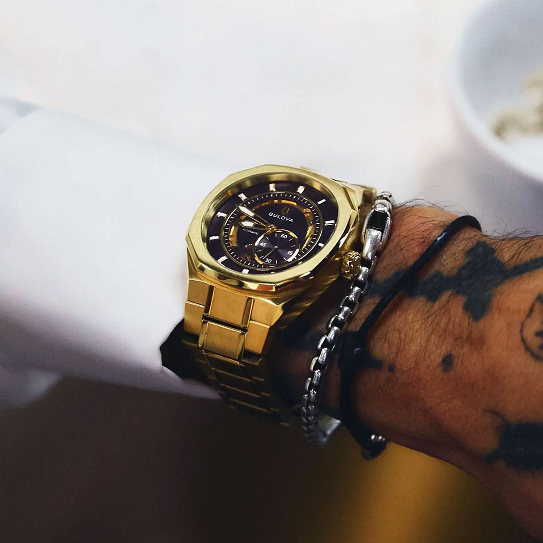 A tattooed wrist displays a Bulova Surveyor Marc Anthony Automatic 40.5mm gold-tone stainless steel men's watch alongside two bracelets—one black, one silver—set against a blurred background.