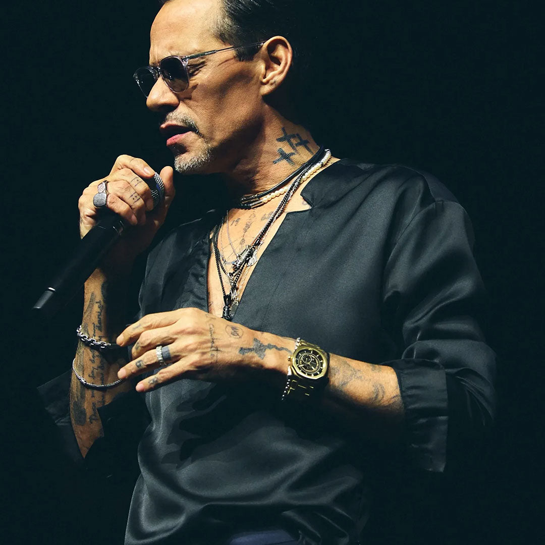A man with tattoos and sunglasses sings into a microphone, wearing a black shirt, necklaces, rings, and the Bulova Surveyor Marc Anthony Automatic 40.5mm Watch by Bulova, featuring gold-tone stainless steel and automatic movement, against a dark background.
