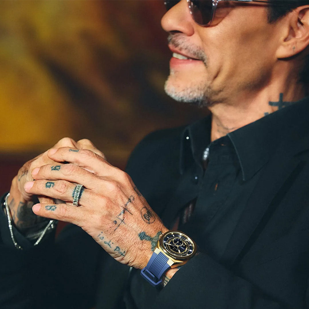 A man with tattoos on his hands wears sunglasses, a black shirt, and the Bulova Surveyor Marc Anthony Automatic 40.5mm Watch, featuring a gold-tone stainless steel case and automatic movement, as he sits with his fingers interlaced.