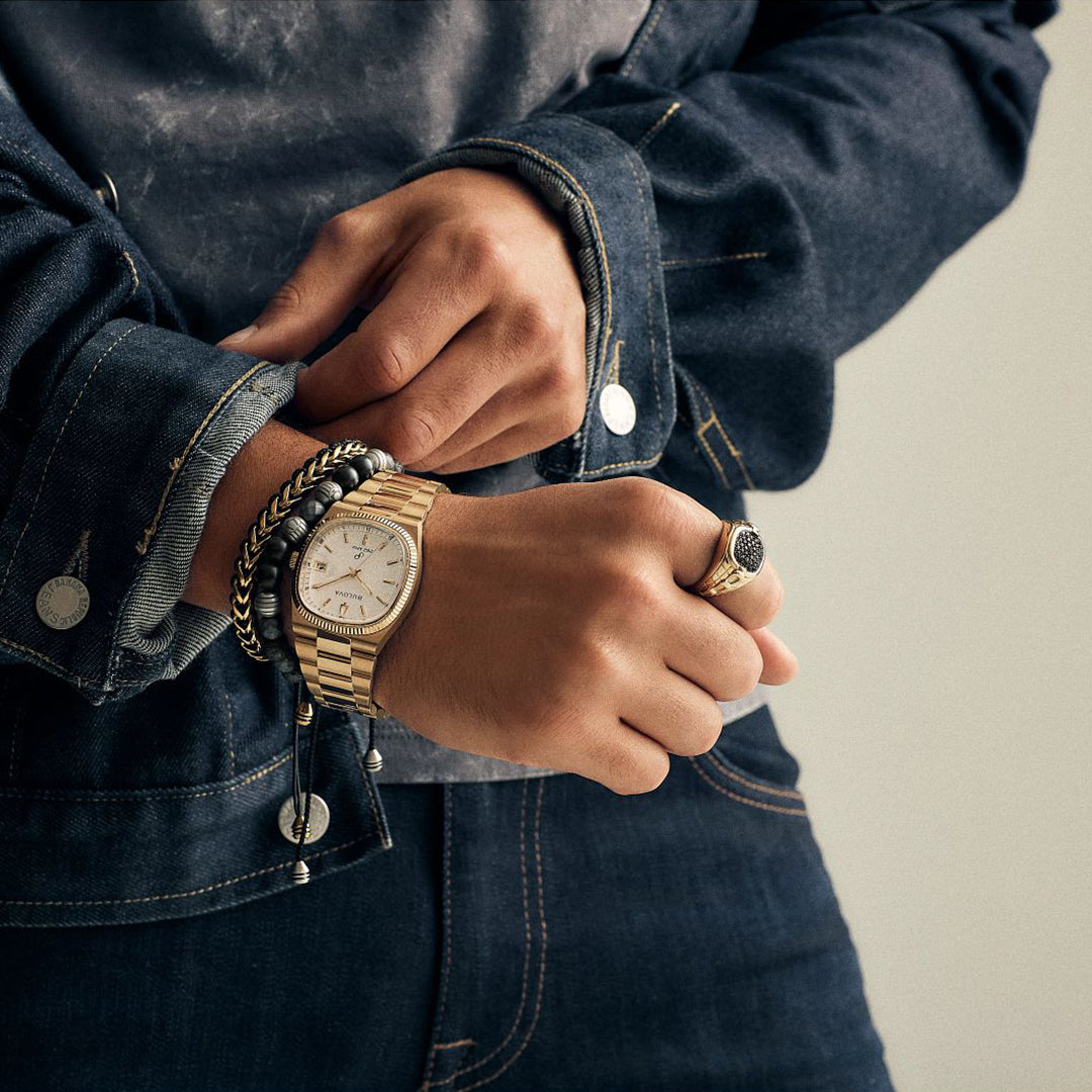 A person wearing a Bulova Super Seville HPQ Precisionist Quartz watch, a black beaded bracelet, and a ring with a dark stone adjusts the cuff of their denim jacket, highlighting the accessories and clothing details.