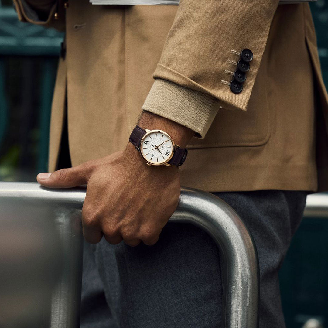 Wearing a tan blazer and gray pants, a person leans on a metal railing, their wrist graced by the sophisticated Bulova Hudson Automatic 39mm Watch. This timepiece from Bulova captures timeless elegance with its automatic movement, white face adorned with Roman numerals, and a brown leather strap.