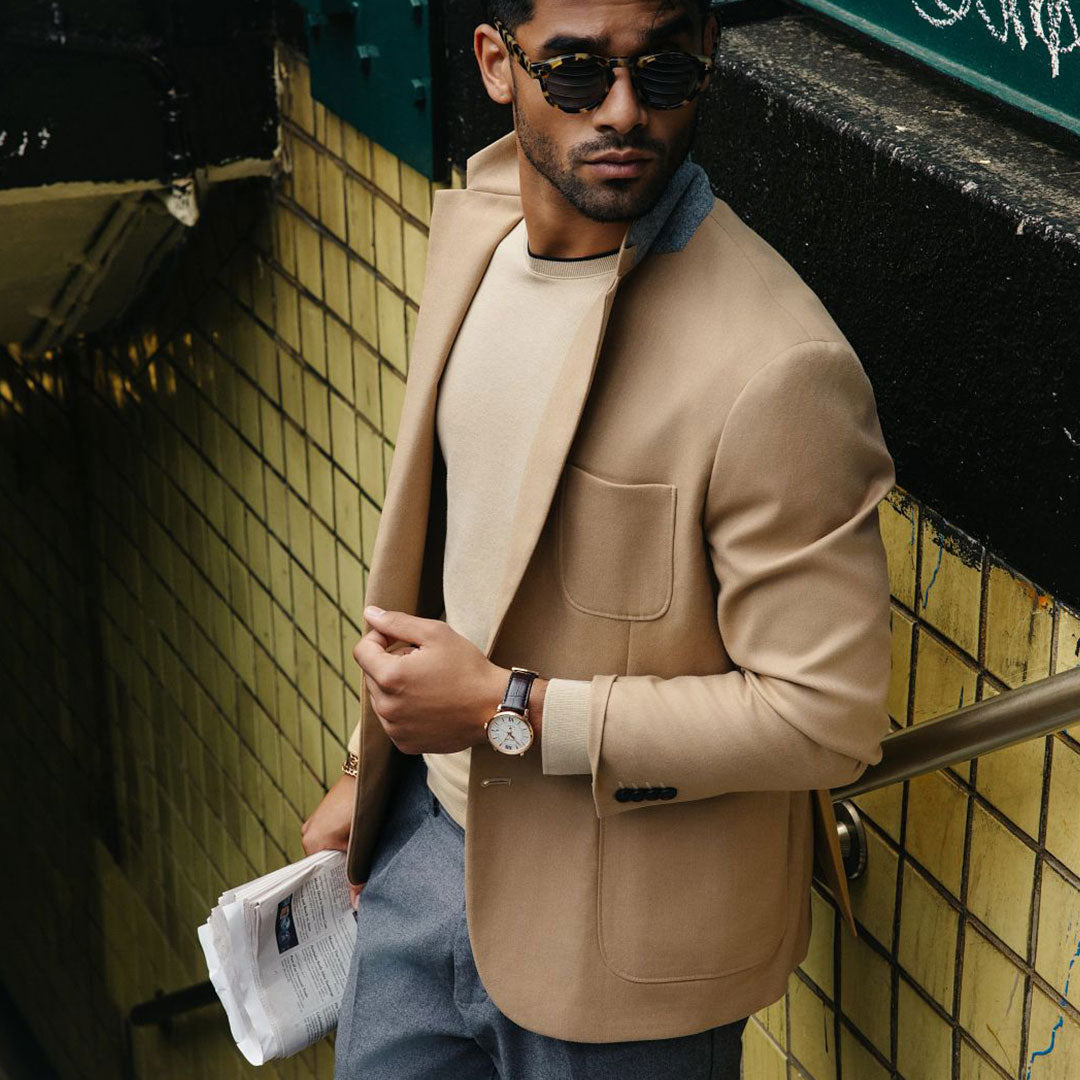 A man with sunglasses, a beige blazer, and a cream shirt stands on a staircase holding a newspaper. On his left wrist is a rose gold-tone stainless steel Bulova Hudson Automatic 39mm Watch. He gazes to the side, with tiled walls forming the backdrop.