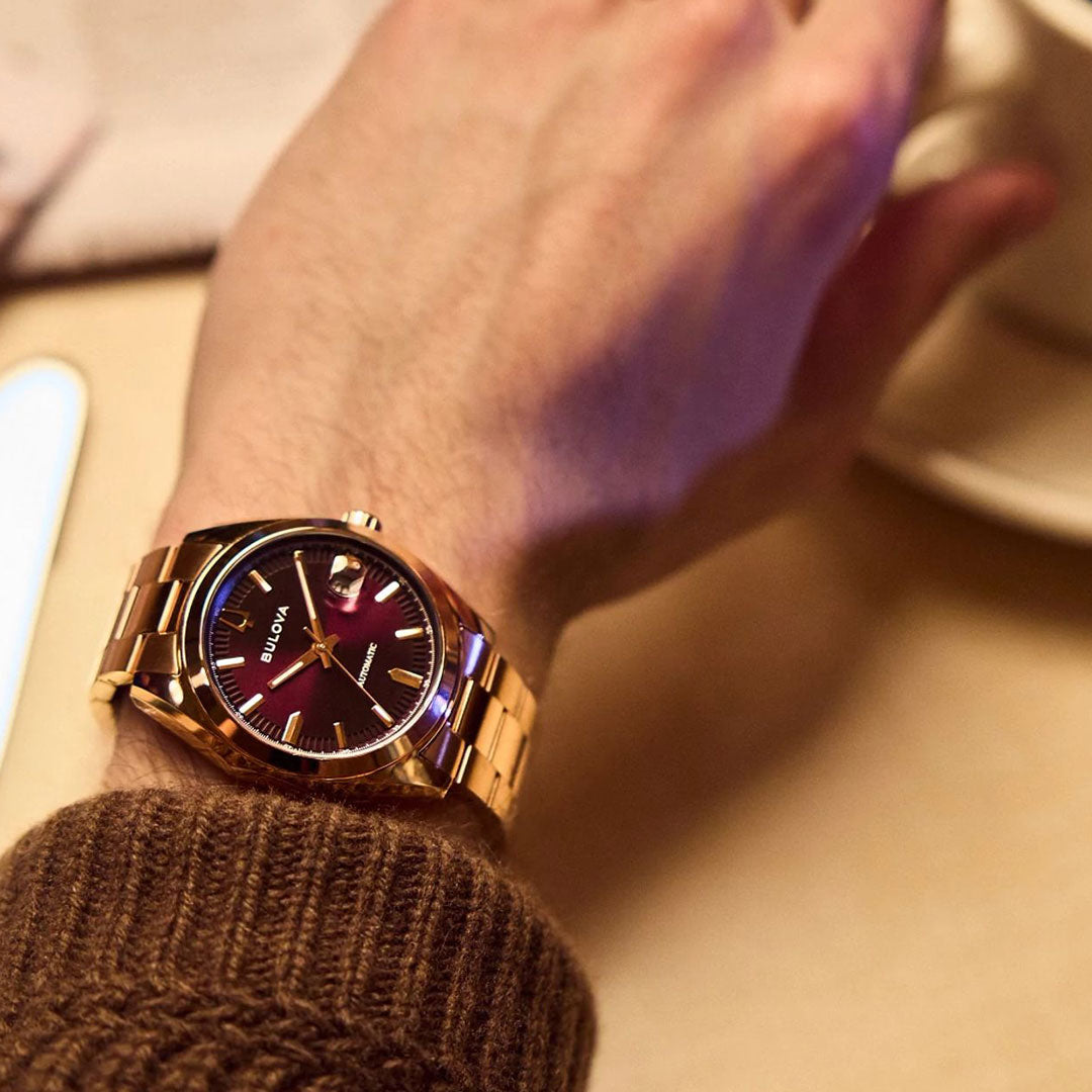 A person wears the Bulova Surveyor Automatic 39mm Watch, featuring a black dial and gold markers, holding a white cup. The rose gold-tone Bulova timepiece contrasts with their brown knit sweater.