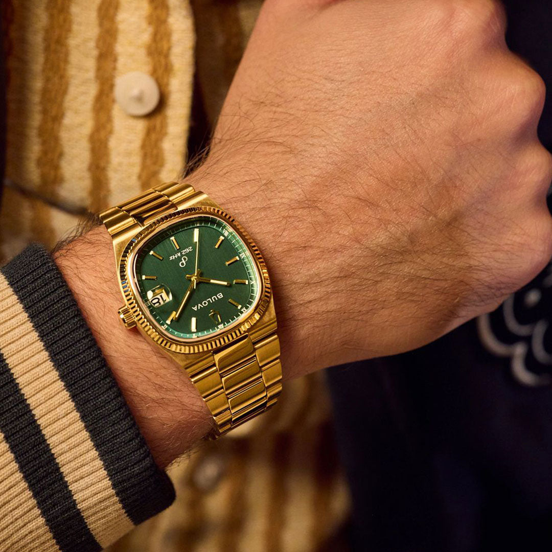 A person wears a Bulova Super Seville HPQ Precisionist Quartz 37.5mm men's gold-tone watch with a green face and gold band, paired with a striped knit sweater. The watch displays the date and time.