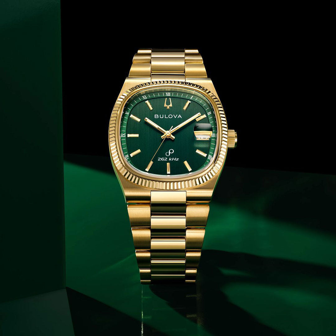 A Bulova Super Seville HPQ Precisionist Quartz 37.5mm watch with a green dial, gold markers, and matching metal band is displayed against a dark green and black background, with the date at 3 o’clock.
