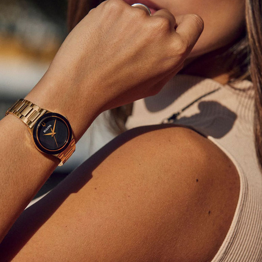 A person wearing a chic Bulova Millennia Quartz 32mm watch, featuring a black circular face and slender orange hands, paired with a sleeveless beige top. The gold-tone stainless steel of the Bulova watch shines beautifully in the sunlight, highlighting its sophisticated and contemporary design.