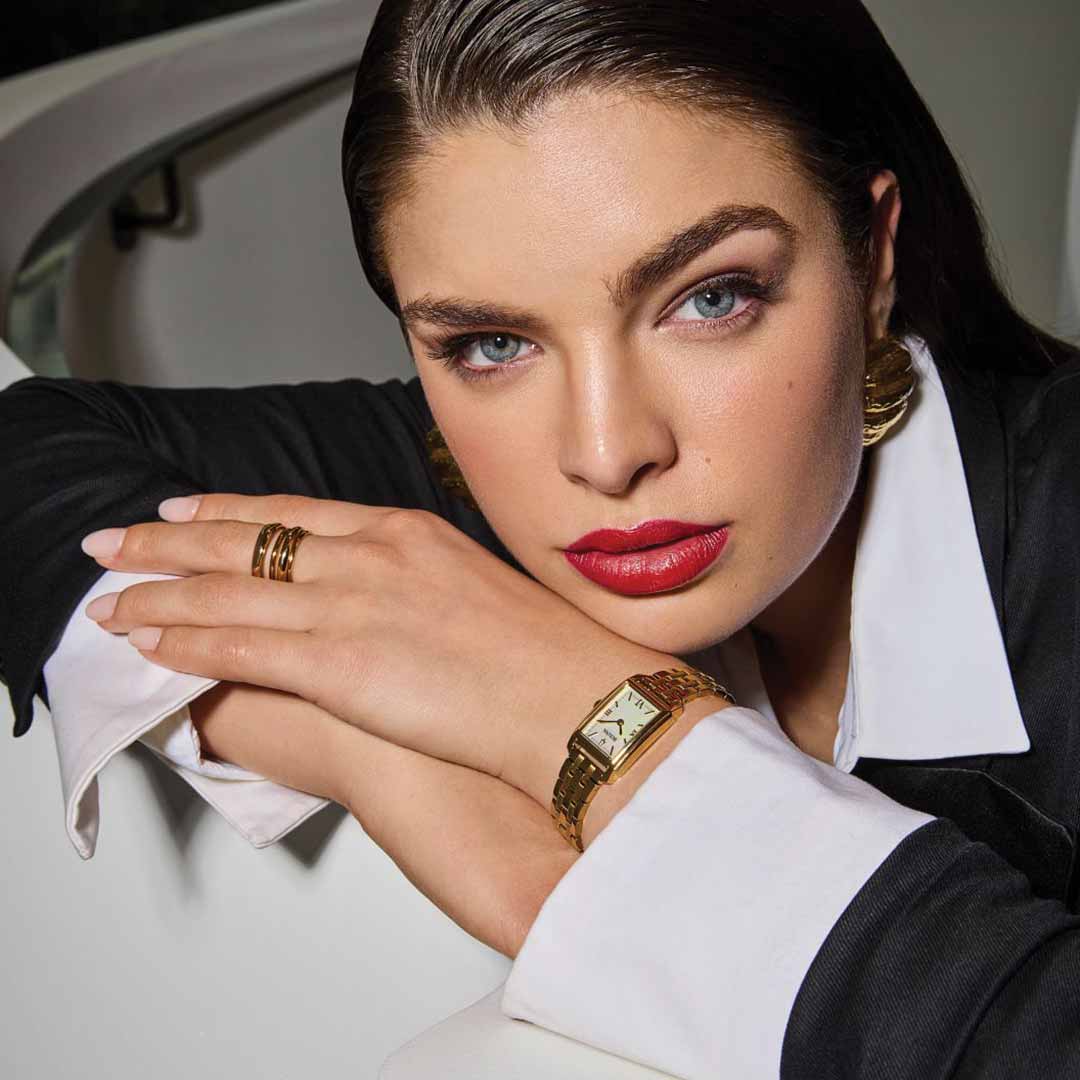 A person with long hair and bold red lipstick leans on a white surface, showcasing a black and white outfit, gold earrings, and the Bulova Sutton Quartz 21mm women's timepiece in gold-tone stainless steel. Their look is completed by a watch featuring a mother-of-pearl dial as they stand before a modern, curved white staircase.