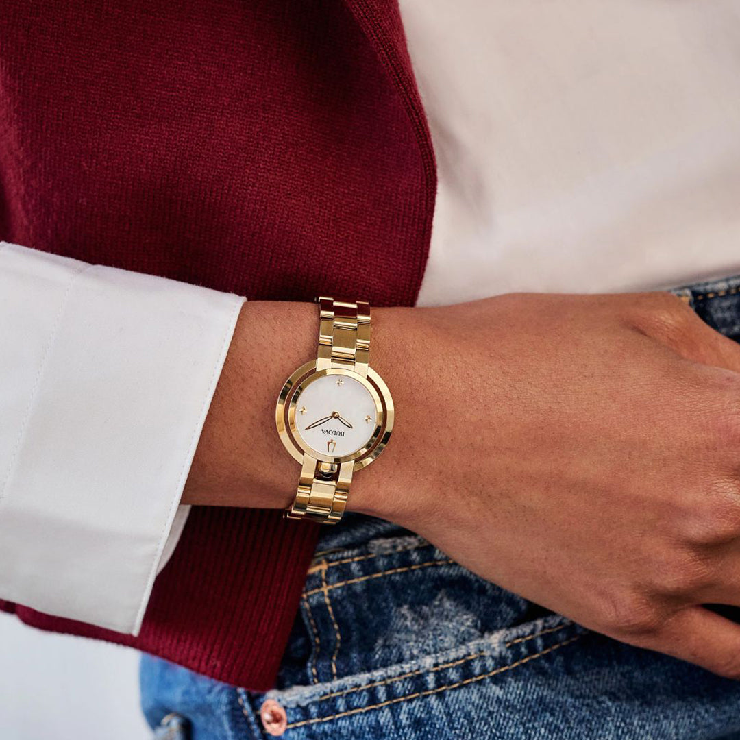 A person is elegantly dressed in a white shirt, red jacket, and blue jeans. Their hand, adorned with a Bulova Rubaiyat Quartz 30.5mm Watch featuring a gold-tone stainless steel bracelet and a white dial with gold markers, rests gracefully against their hip.