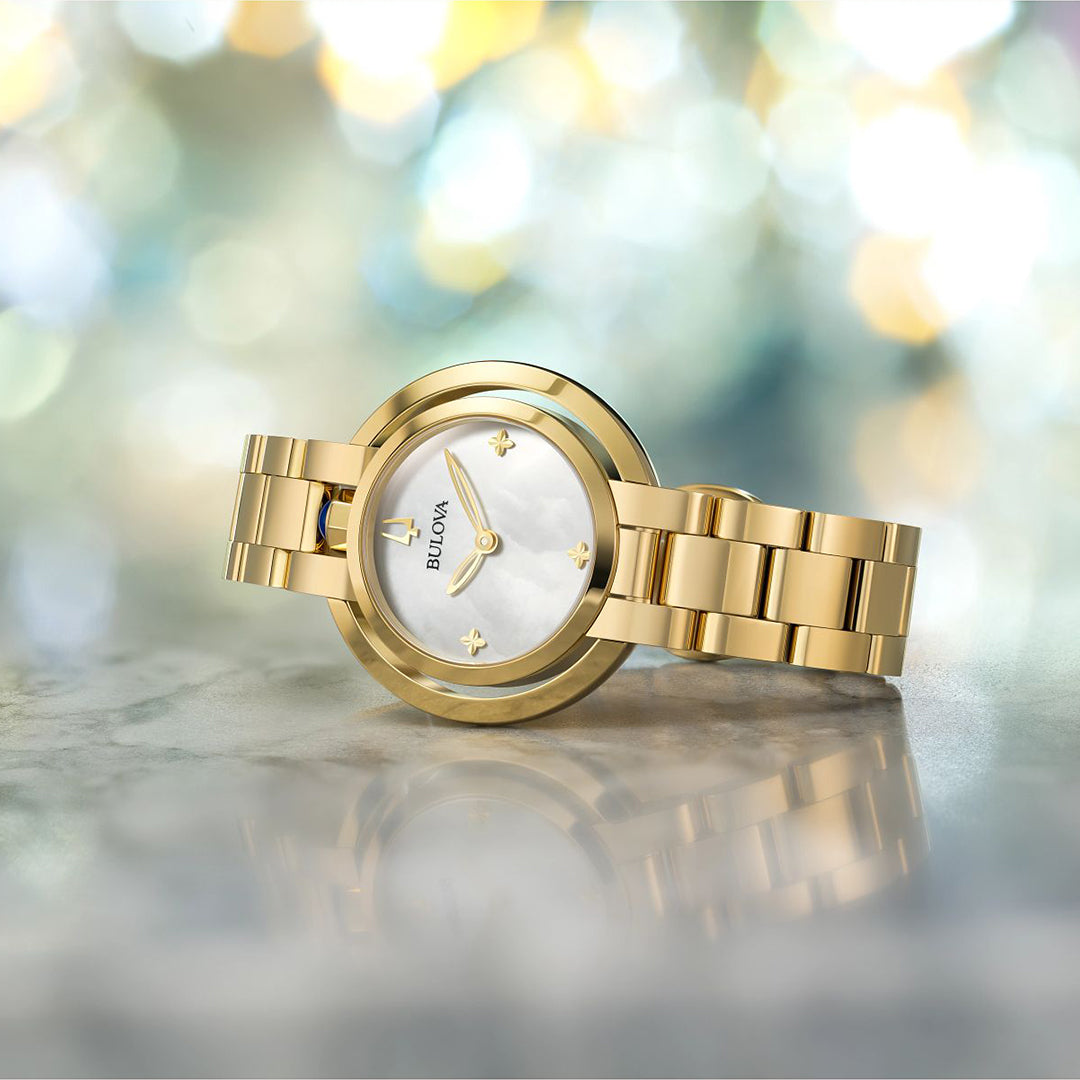 A Bulova Rubaiyat Quartz 30.5mm Watch rests elegantly on marble, its white face and minimalist design enhanced by a gold-tone stainless steel band and a colorful bokeh background, proudly displaying the Bulova name.