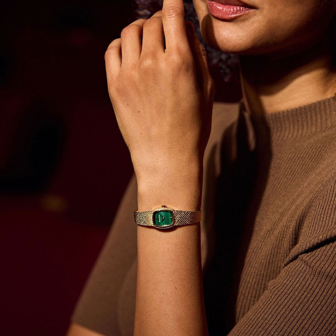 A woman in a brown short-sleeve knit top showcases the Bulova Goddess of Time Quartz 17mm Watch, featuring a green rectangular face and gold-tone stainless steel, with her hand near her chin against a softly blurred background.