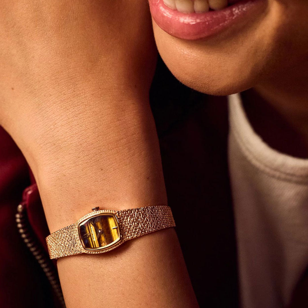 A close-up of a person smiling, wearing the Bulova Goddess of Time Quartz 17mm watch with a rectangular yellow face and textured gold band by Bulova on their wrist.