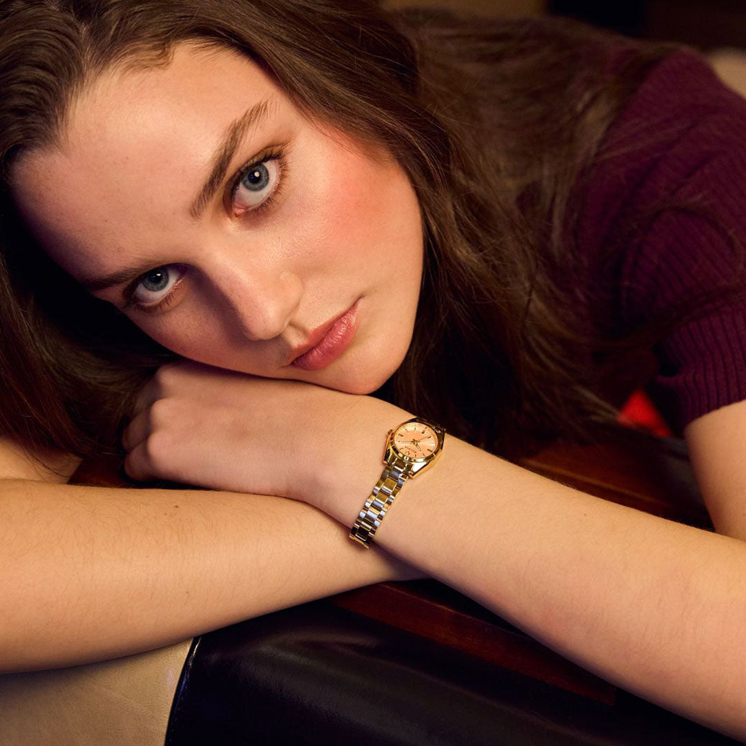 A young woman with long brown hair, wearing a burgundy top and a Bulova Surveyor Quartz 23mm Watch, rests her head and arms on a table, gazing at the camera under warm, soft lighting.