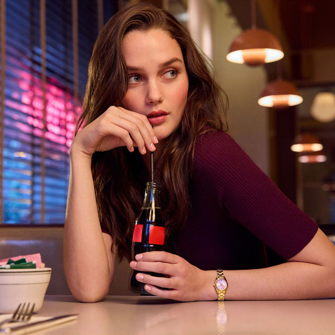A woman with long brown hair in a maroon top sits at a diner table with neon lights, sipping soda and wearing the Bulova Surveyor Quartz 23mm Watch by Bulova, her gaze directed pensively to the side.