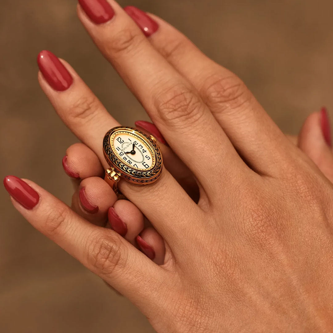 A hand with red-painted nails wears a Bulova Ring Quartz 17mm Watch, featuring a small oval face showing 11:55 in vintage gold-tone, set against a brown background.