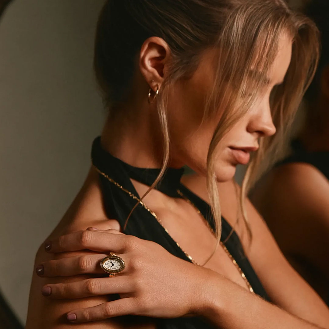 A woman with blonde hair in a loose updo poses in profile, accessorized with a black halter top, gold hoop earrings and necklace, and the Bulova Ring Quartz 17mm Watch by Bulova featuring a round decorative design on her hand.