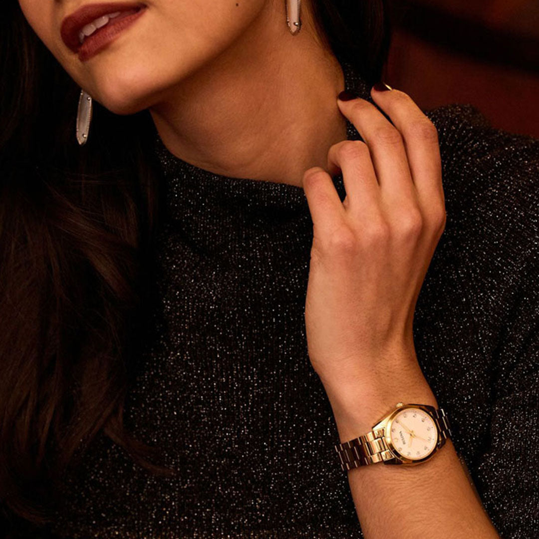 A woman wearing a sparkling black turtleneck sweater and large earrings rests her hand on her neck, showcasing a classic Bulova Surveyor Quartz 31mm watch with a gold-tone stainless steel band on her wrist. Her dark hair is partially visible, adding to the elegant allure of her ensemble.