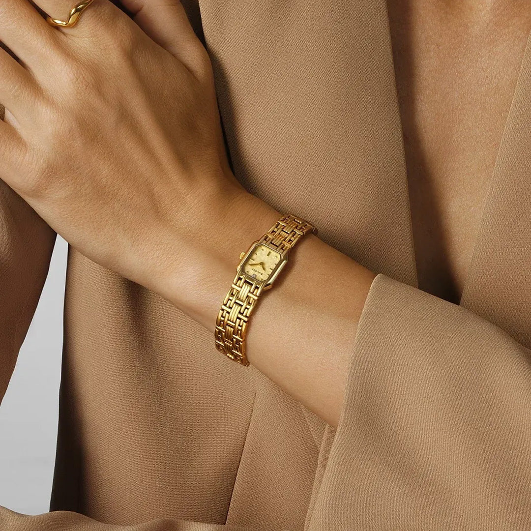 A person wearing the Bulova Goddess of Time Sophia Quartz 16mm Watch, featuring a gold-tone stainless steel band and a diamond accent, along with a gold ring, rests their hand on their chest. They are dressed in a light brown suit jacket.