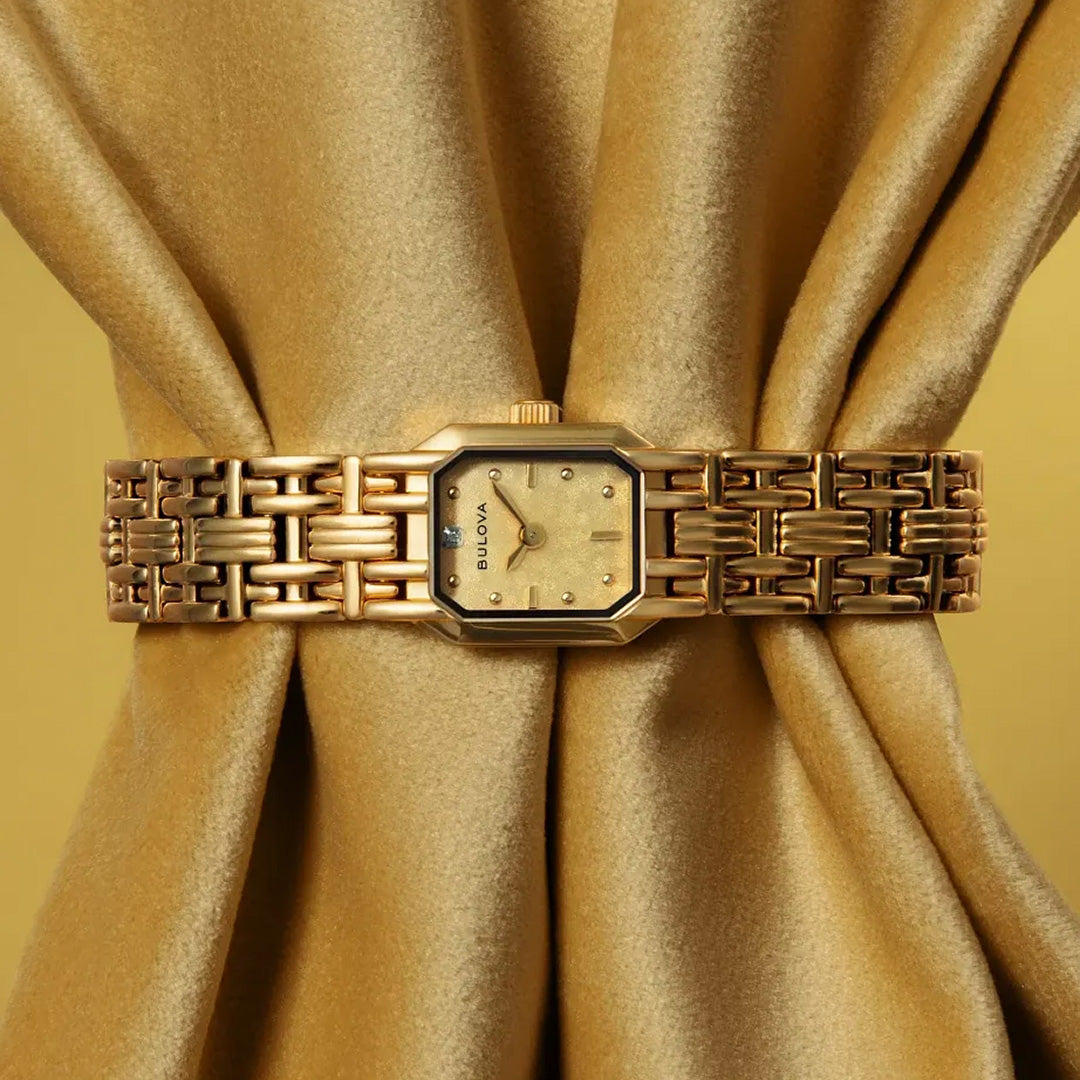 A gold-tone stainless steel Bulova Goddess of Time Sophia Quartz 16mm Watch with a geometric band is elegantly wrapped around shimmering gold fabric, gathered at the center to create a luxurious display. Subtle diamond accents on this stunning Bulova timepiece enhance its sophisticated and refined appeal.