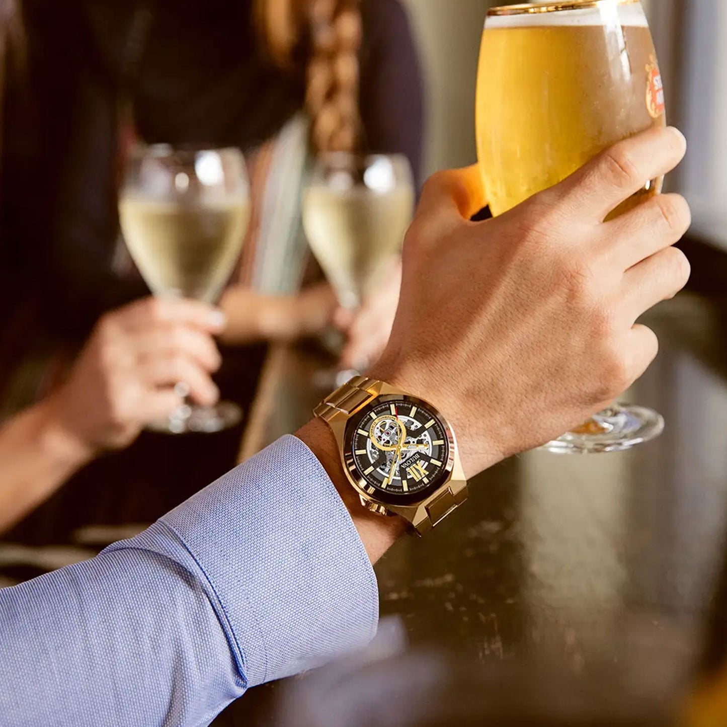 A person wearing a Bulova Maquina Automatic 46mm gold-tone stainless steel watch holds a pint of beer. In the background, two people hold glasses filled with white wine, suggesting the scene is set in a casual environment, possibly a bar or restaurant.