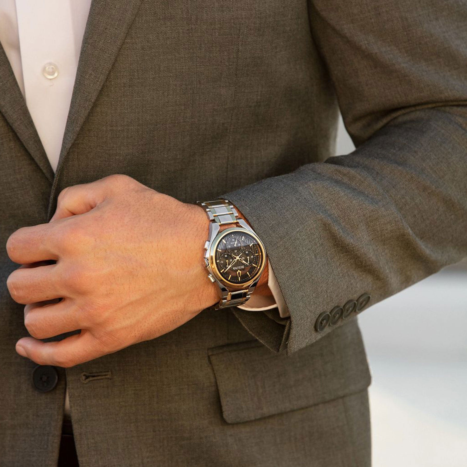 A person in a gray suit holds their wrist to showcase an elegant Bulova CURV HPQ Quartz 44mm watch with a dark face and metallic strap. The focus is on the suit's detail and the watch's intricate curved chronograph movement.