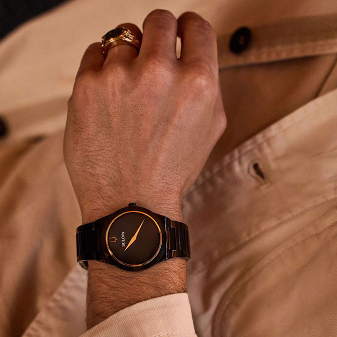 A person wearing a Bulova Millennia Black Quartz 41mm Watch rests their hand on a beige shirt. The sleek black watch, with its minimalist design featuring a round face and gold accents, complements the gold ring on their index finger.