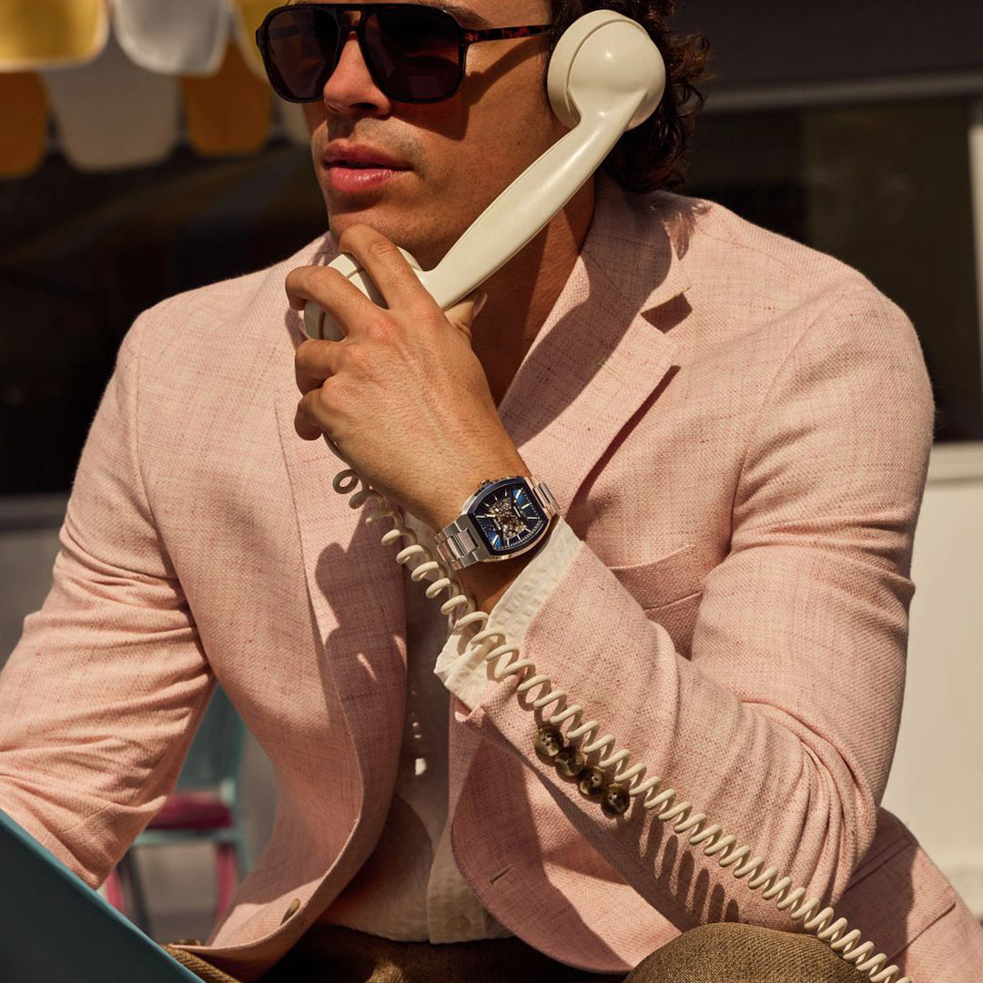 A man in a light pink blazer and sunglasses talks on a vintage corded phone, wearing the Bulova Maquina Automatic 40mm Watch by Bulova, as sunlight casts stylish shadows on his face.