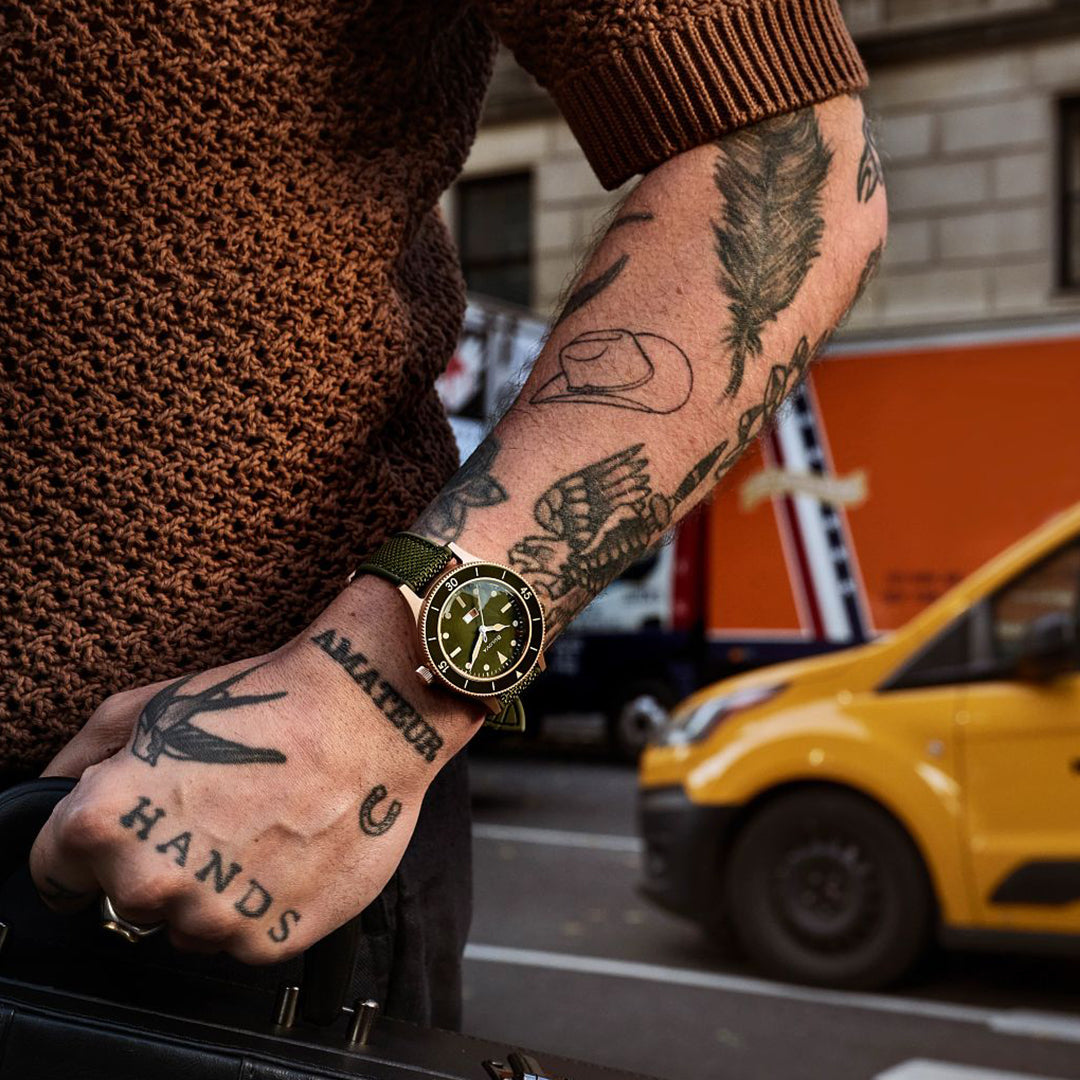 A person with tattoos like "Amateur" and "Hands" wears a Bulova Mil Ships Automatic 41mm Watch, marking the brand's 150th Anniversary. They're holding a black bag, while a yellow van and an orange truck navigate the bustling city street behind them.