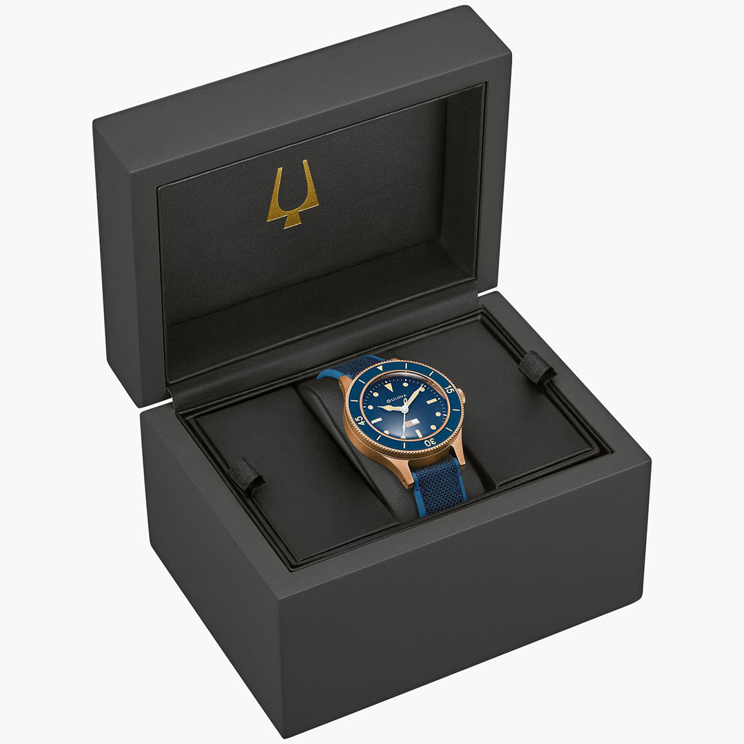 The Bulova MIL Ships CUSN8 Bronze Archive Series Automatic 41mm watch is shown in an open black box with a gold trident logo on the lid. The watch features a blue and gold design with a blue strap.