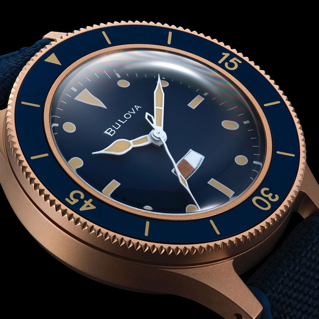 Close-up of the Bulova MIL Ships CUSN8 Bronze Archive Series Automatic 41mm Watch, featuring a dark blue dial with gold hands and markers, a CuSn8 bronze case, blue and gold bezel, and a dark textured strap.