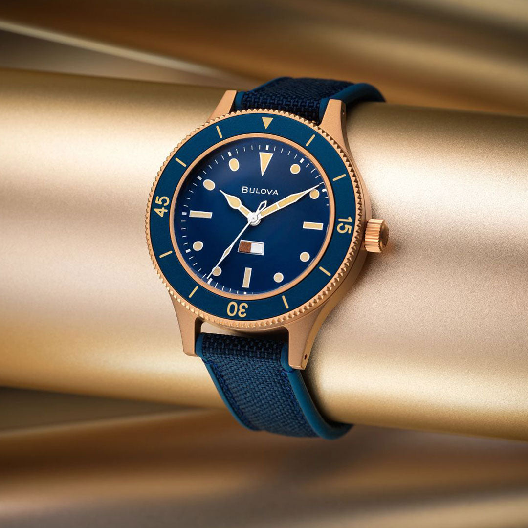 The Bulova MIL Ships CUSN8 Bronze Archive Series Automatic 41mm Watch, featuring a blue dial and fabric strap, is displayed wrapped around a gold metallic cylinder with a blurred gold background.