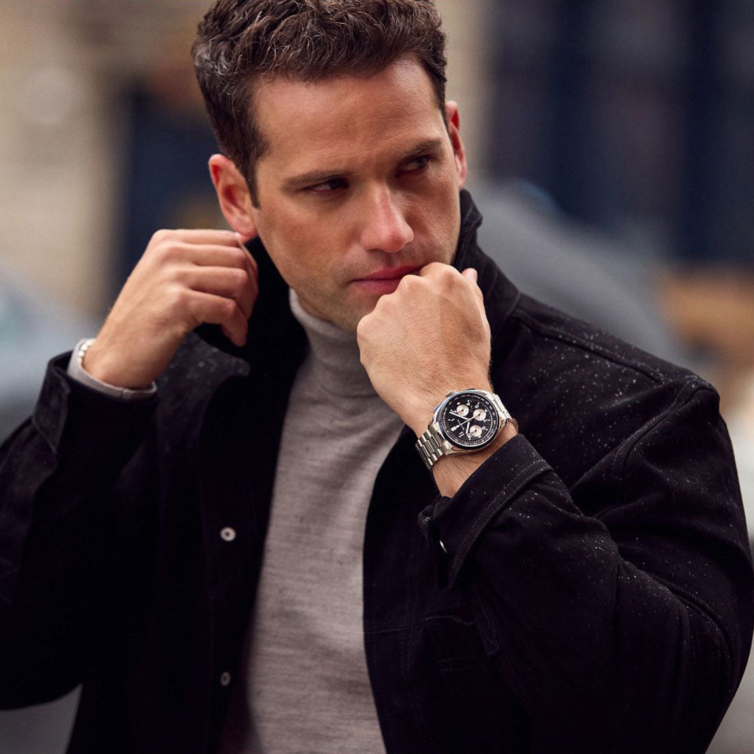 A man in a light gray turtleneck and black jacket adjusts his collar while glancing sideways, wearing a Bulova Lunar Pilot Timascus Quartz 43.5mm Limited Edition watch on his left wrist. The softly blurred background enhances the scene.