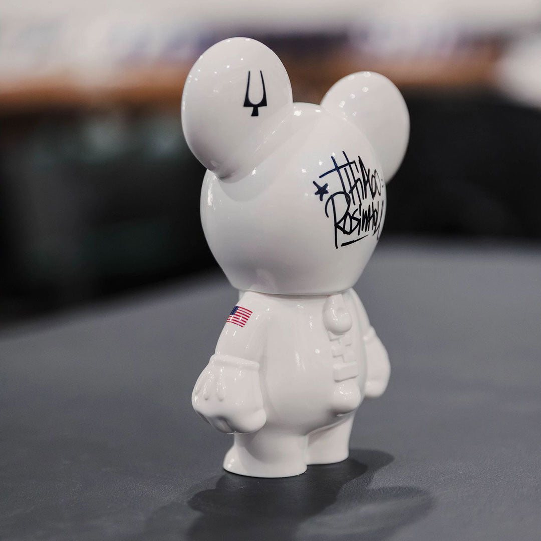 A white, mouse-shaped astronaut figurine stands on a table, sporting an American flag patch, trident symbol, and black graffiti-style text—its suit inspired by the Bulova Lunar Pilot Timascus Quartz 43.5mm Watch Limited Edition.