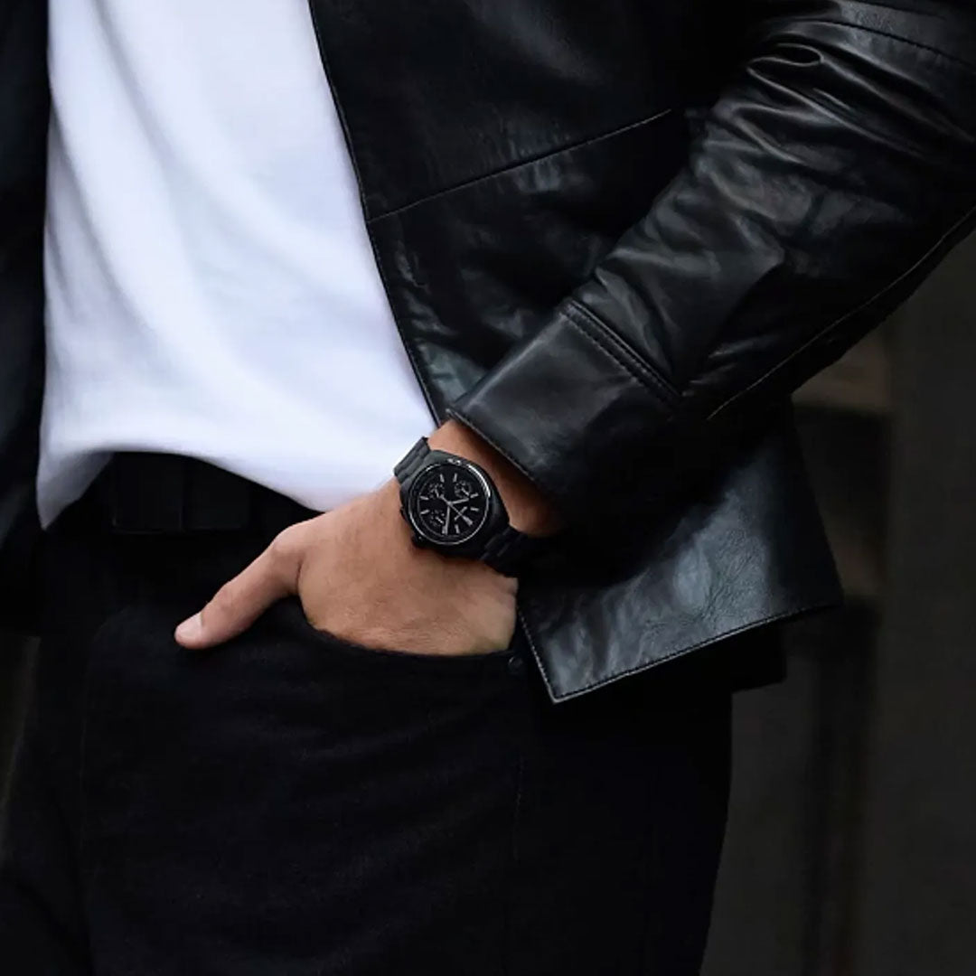 A person wearing a black leather jacket, white shirt, and black pants has one hand in their pocket and is sporting the Bulova Archive Series Luna Pilot Black Hole Quartz 41.2mm watch, renowned for its high precision quartz movement.