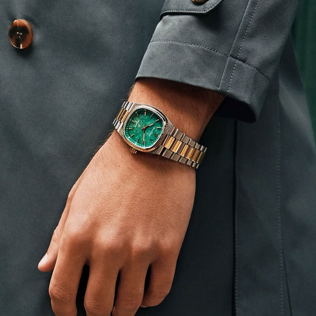 A person wearing a gray coat and a Bulova Super Seville HPQ Precisionist Quartz 35mm men's luxury watch with a gold and silver band and green face poses with their hand relaxed and visible from the wrist down.
