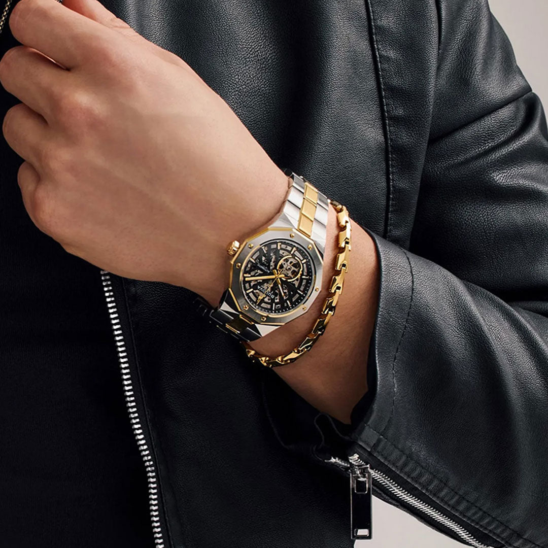 A person wearing a black leather jacket showcases the Bulova Prestige Automatic 42mm Watch, which features a silver and gold case, a black skeleton dial beneath sapphire crystal, and a gold chain bracelet on their wrist.