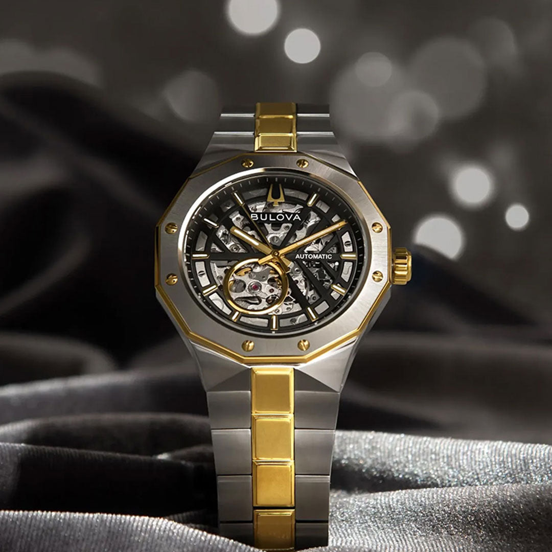 The Bulova Prestige Automatic 42mm Watch by Bulova, featuring a gold and silver-tone band, rests on gray fabric. This timepiece showcases automatic movement, an open skeleton dial with visible gears and gold accents, set against a softly blurred bokeh background.