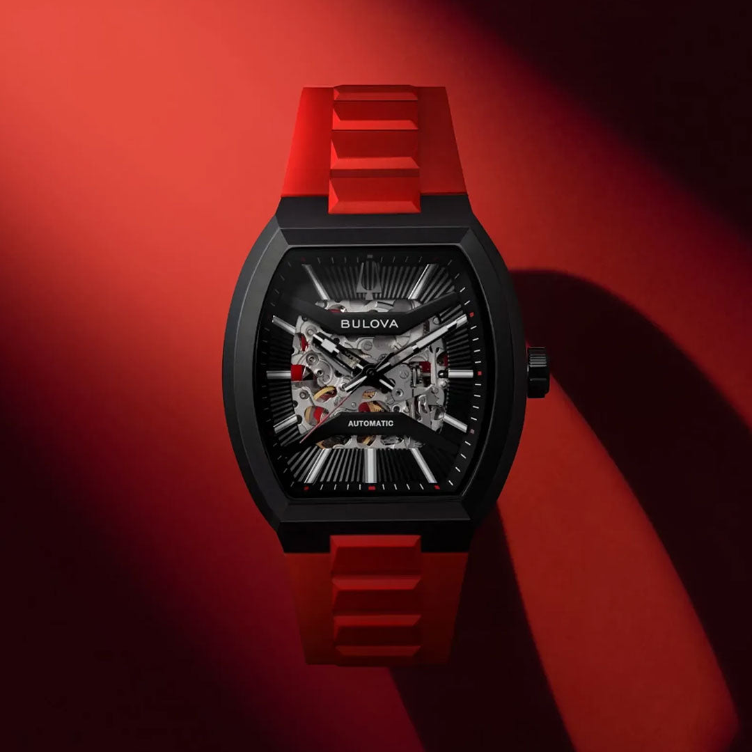 A Bulova Maquina Automatic 40mm Watch by Bulova, featuring a striking red strap and black-toned case with an automatic skeleton dial that showcases its inner workings, is presented against a bold red and black gradient background highlighted by dramatic lighting and shadows.