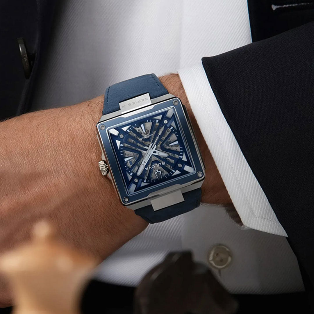 Wearing a dark suit, an individual sports the Bulova Series X Automatic 41mm Watch—a square, blue-faced timepiece with a matching blue strap and exposed gears that highlight its skeletonized automatic movement—resting their arm on a table scattered with chess pieces in the foreground, capturing a scene that embodies the refined artistry of Bulova luxury watchmaking.