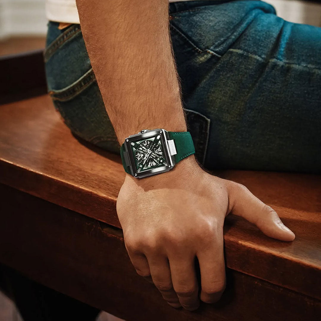 A person wearing blue jeans sits on a wooden surface, resting their left arm down. They are wearing a Bulova Series X Automatic 41mm Watch—a sophisticated timepiece with a square case, green strap, and a black, intricately detailed watch face—an outstanding example of luxury craftsmanship from Bulova.