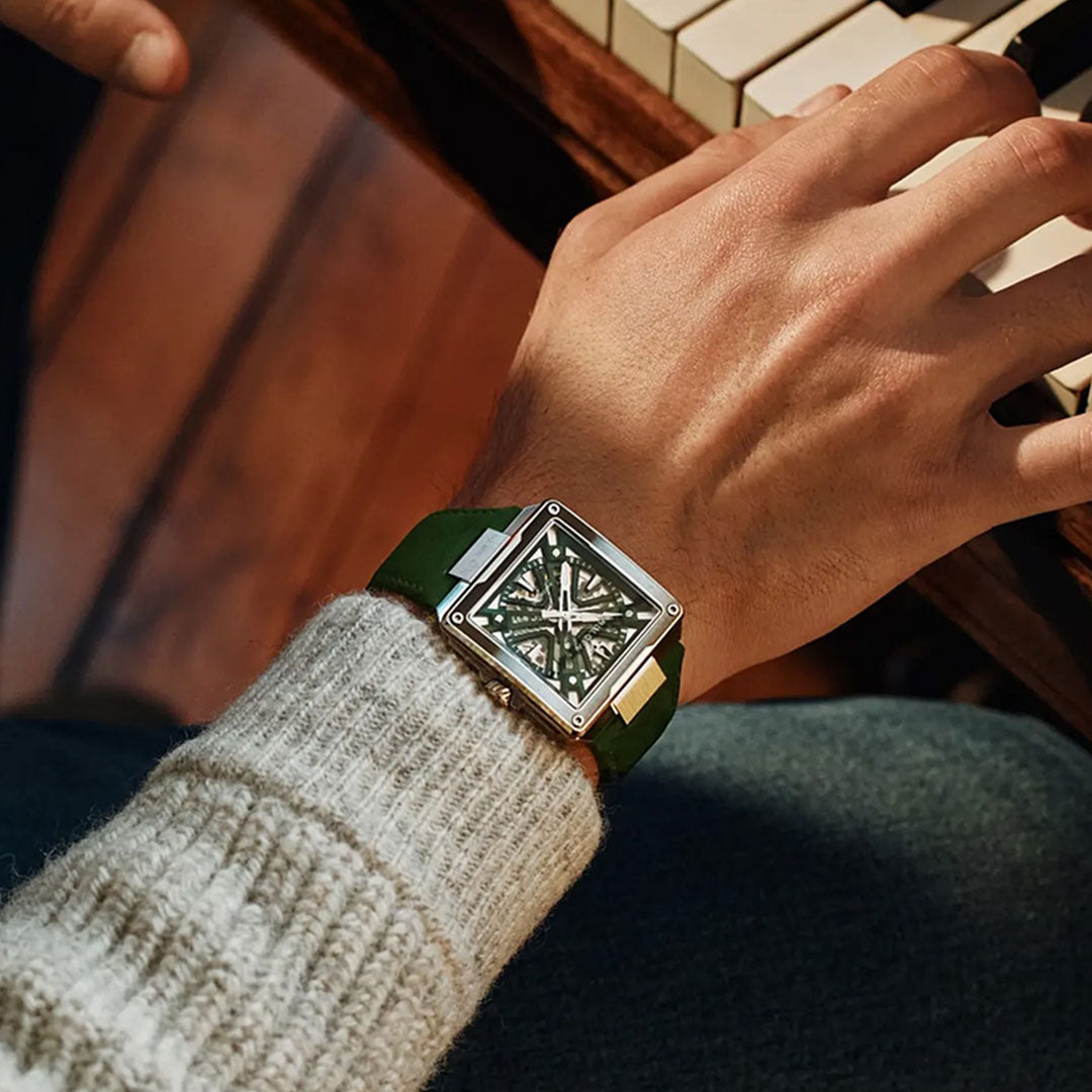 A person wearing a green-banded, square-faced Bulova Series X Automatic 41mm Watch plays piano. Only their hand, wrist, and the sleeve of their sweater are visible alongside piano keys and a wooden floor in the background—a subtle showcase of Bulova’s luxury watchmaking craftsmanship.