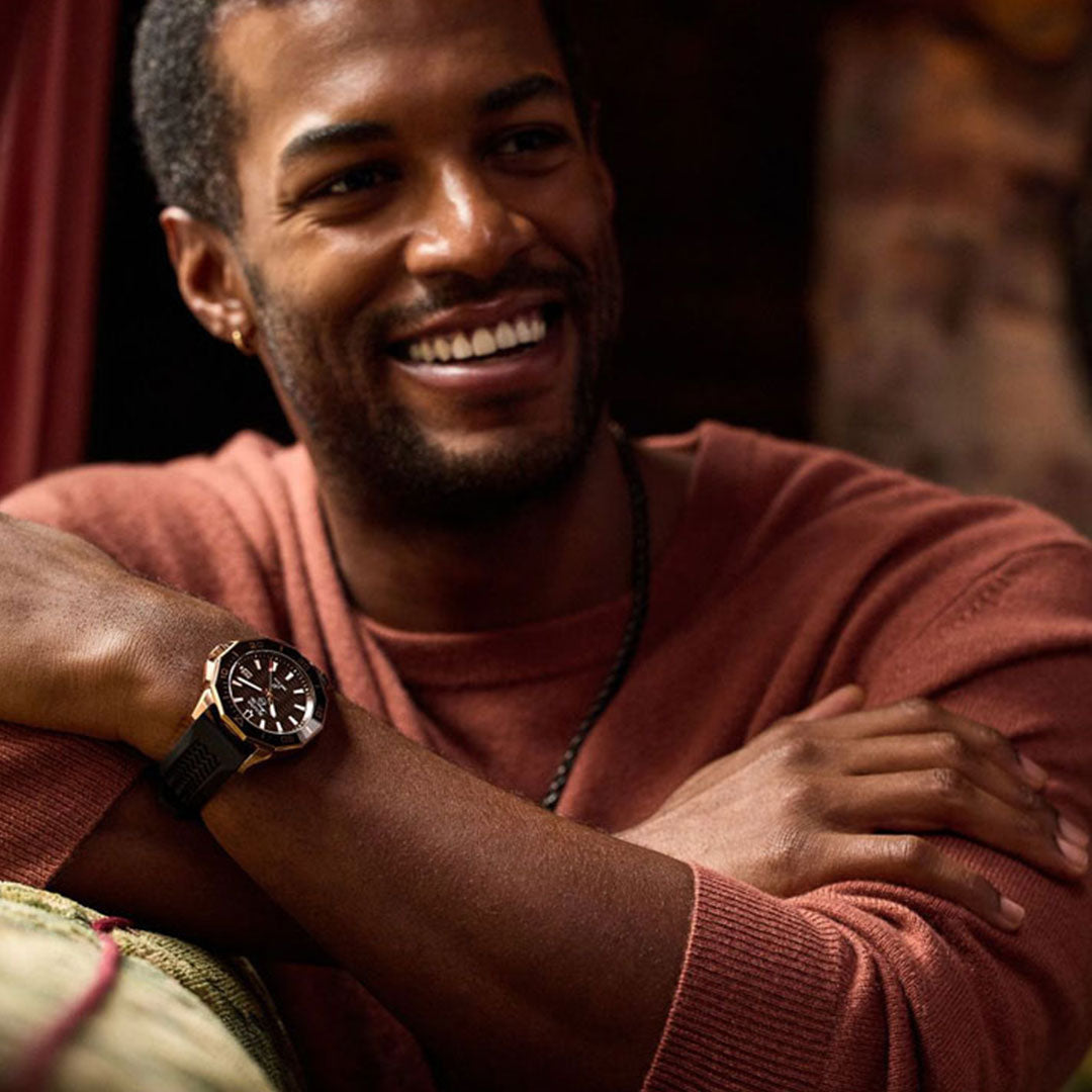 In a comfortable setting, a man smiles warmly while wearing a brown sweater and proudly displaying a Bulova Marine Star Quartz 43mm Watch with rose gold-tone stainless steel on his wrist. The blurred interior background adds to the relaxed and inviting atmosphere.