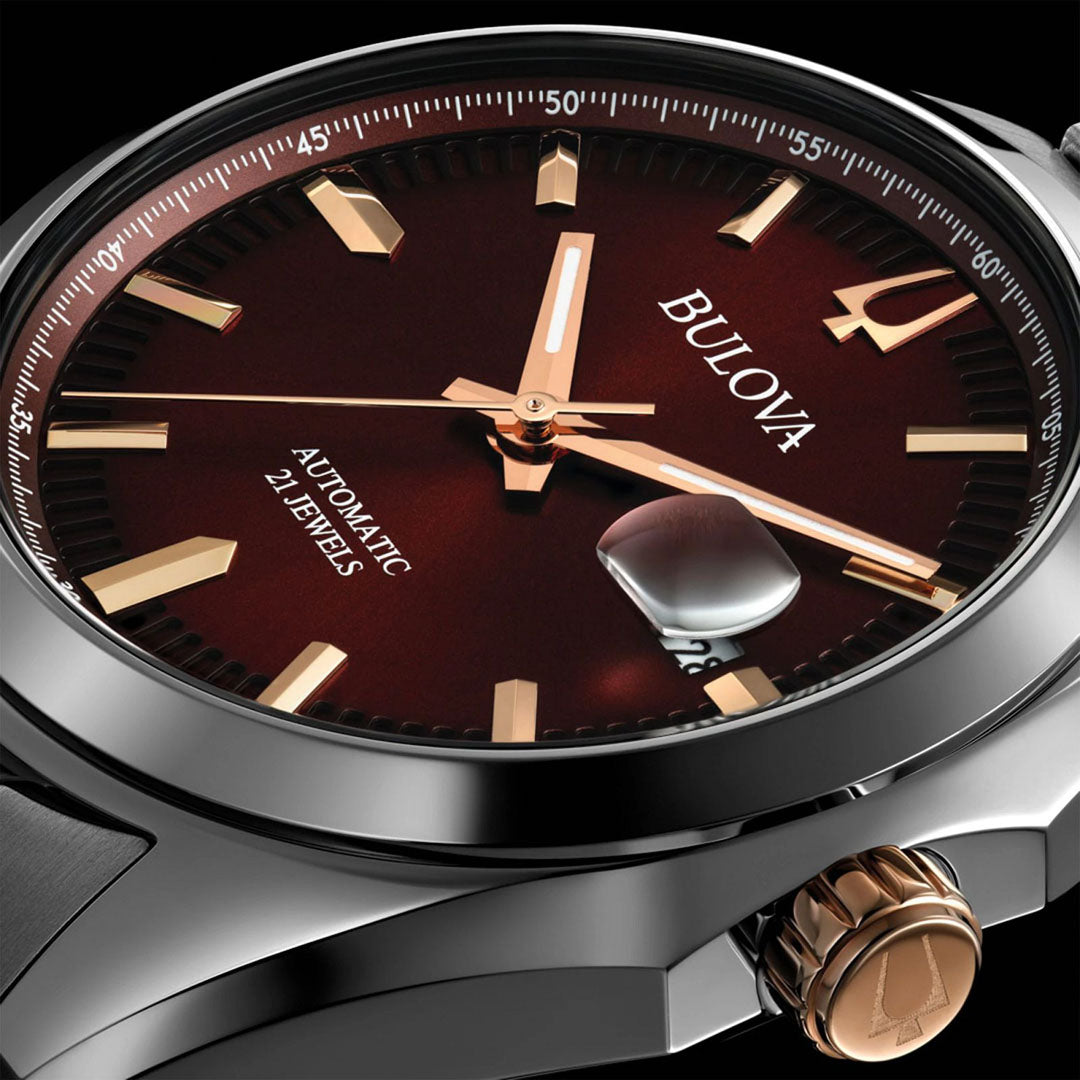 Close-up image of the Bulova Surveyor Automatic 39mm Watch for men, featuring a gleaming polished silver case and a deep burgundy dial. The face showcases gold hour markers, luminous hands, a date window, and the inscription "Automatic 21 Jewels." It includes a textured crown for easy handling and is complemented by an elegant stainless steel bracelet.