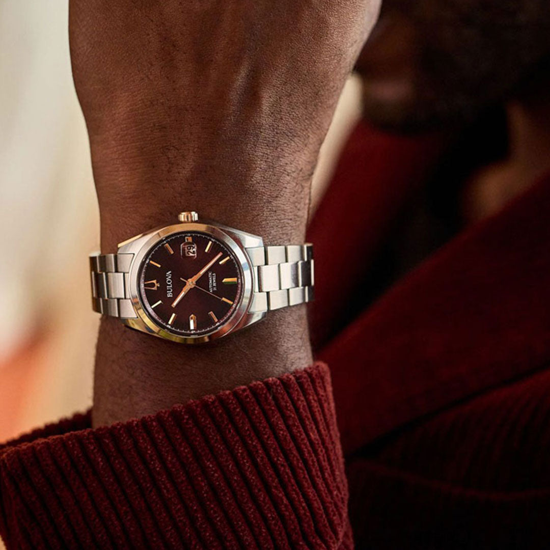 Close-up of a person wearing the stylish Bulova Surveyor Automatic 39mm Watch featuring a dark face and date display, complemented by a stainless steel bracelet. The watch catches the light exquisitely against a maroon ribbed sleeve, making it a refined accessory worn on the left wrist.