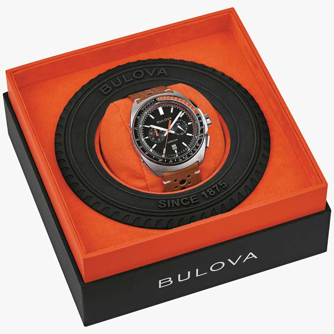 A Bulova Racer Quartz 42mm Watch with a brown leather racing-style strap is showcased in a box featuring an orange and black color scheme. The packaging, embossed with "Bulova Since 1875" and accented by a tire tread design, amplifies its automotive aesthetic.