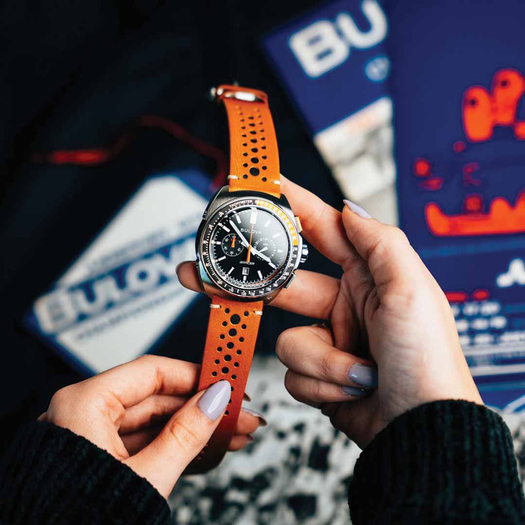 A person with painted nails holds a Bulova Racer Quartz 42mm watch, which showcases a brown racing-style strap and a sleek black dial. In the blurred background, various items and packaging subtly display the Bulova brand name, enhancing the automotive aesthetic.