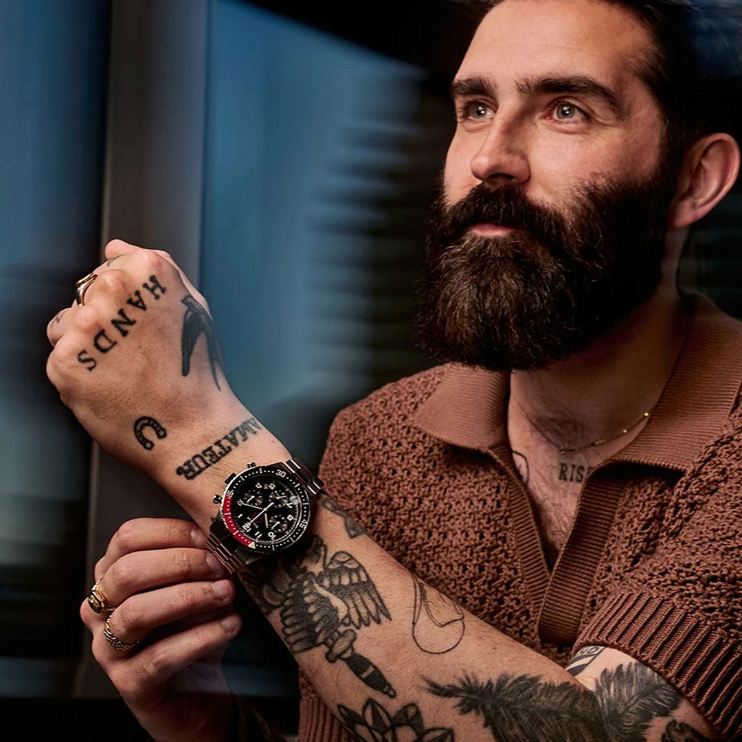 A bearded, tattooed man looks upward, showcasing the Bulova Marine Star Heritage Quartz 42.85mm Watch Limited Edition. He wears a brown knit shirt and gold rings against a softly blurred background.