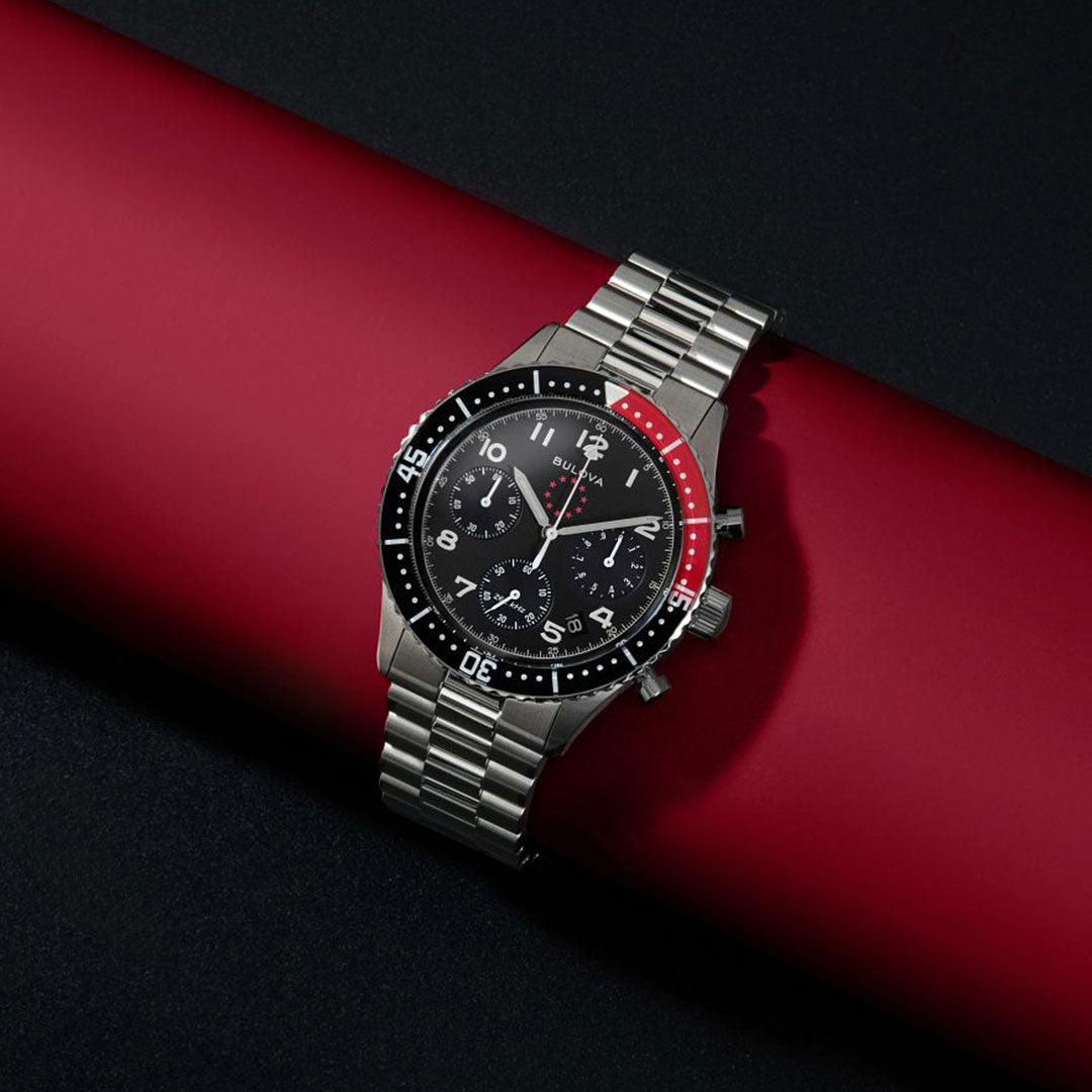 The Bulova Marine Star Heritage Quartz 42.85mm Limited Edition watch features a silver metal band, black and red bezel, and black dial with three subdials, displayed diagonally on a red cylinder, and offers 200m water resistance.