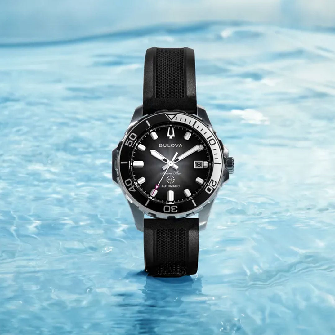A Bulova Marine Star Series B Automatic 41mm watch, in black and silver with a black strap, stands upright on rippling blue water, reflecting light.